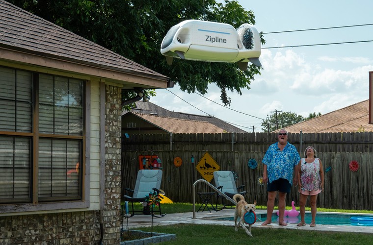A Zipline delivery drone delivers a package to a couple in their backyard.