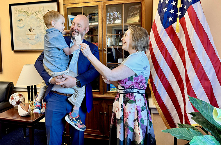 A man is holding a young boy who is giving a woman a photo card. There is an American flag in the background.