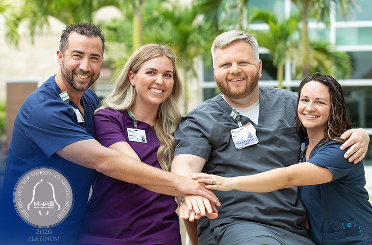Four smiling BayCare team members wearing scrubs stand outdoors with hands stacked in the center, symbolizing teamwork and support. A Bell Seal for Workplace Mental Health Platinum 2026 badge appears in the lower corner of the image.