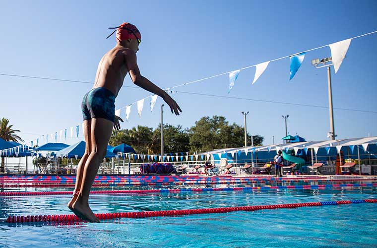A boy wearing a swim cap, goggles and swim trunks jumps into a pool.