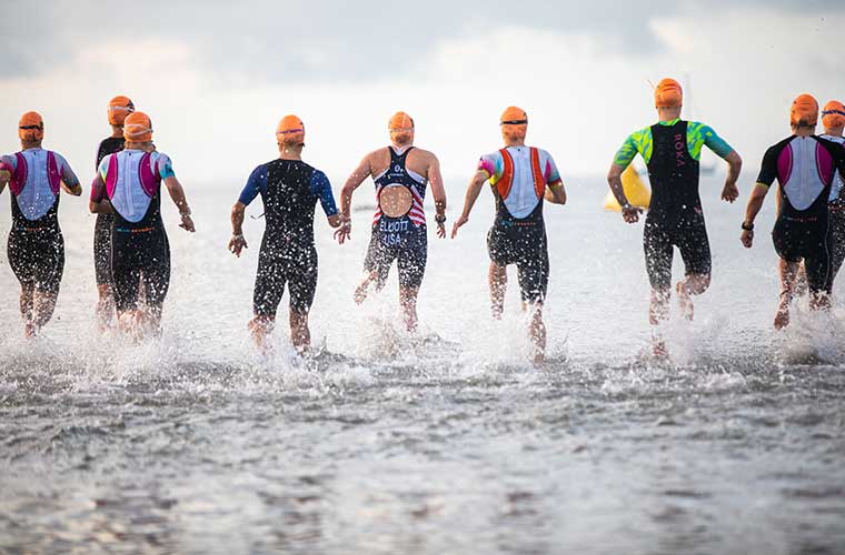 Athletes dressed in swim caps and swimsuits run into the water during a sports event.