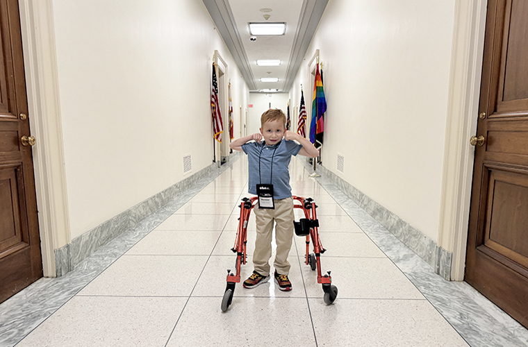 Young boy with a walker standing in a long hallway holding his arms up showing his muscles.