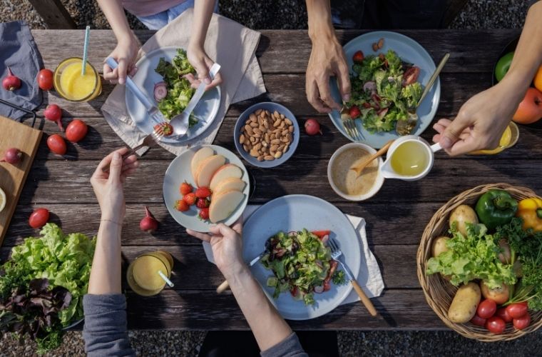 Overhead view of a wooden outdoor table with several plates of fresh salads, sliced fruit and small bowls of dressing, with multiple hands reaching in to serve and eat.