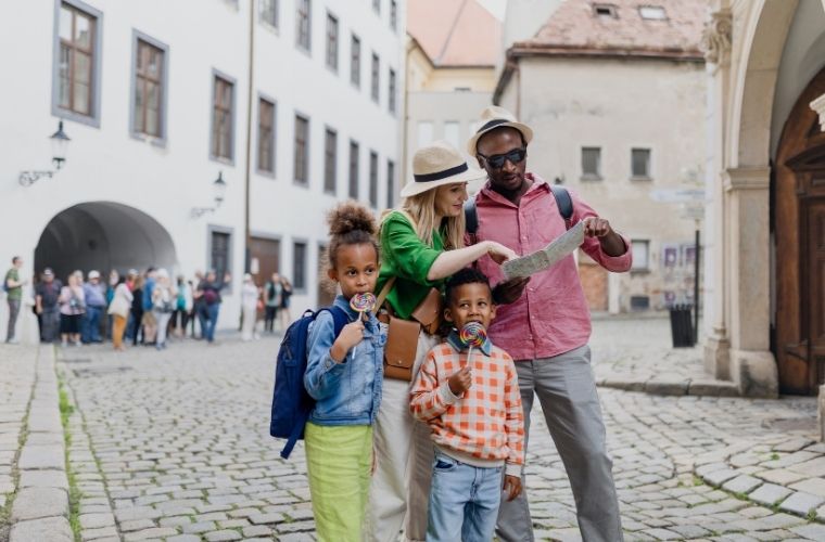 Family of four sightseeing in a historic city, with two adults reviewing a map while two children hold lollipops, on a cobblestone street with old architecture.