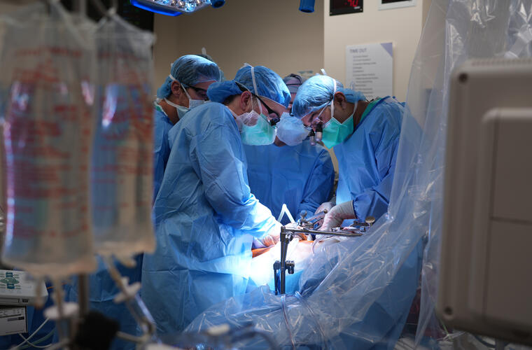 A team of surgeons in blue scrubs and masks performs a complex procedure under bright operating room lights, surrounded by medical equipment.