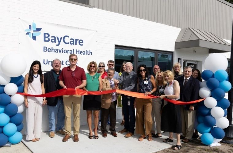 A group of people stands in front of a building, holding a red ribbon for a ceremonial cutting. Behind them is a sign reading "BayCare Behavioral Health Urgent Care." Blue and white balloons decorate the area.