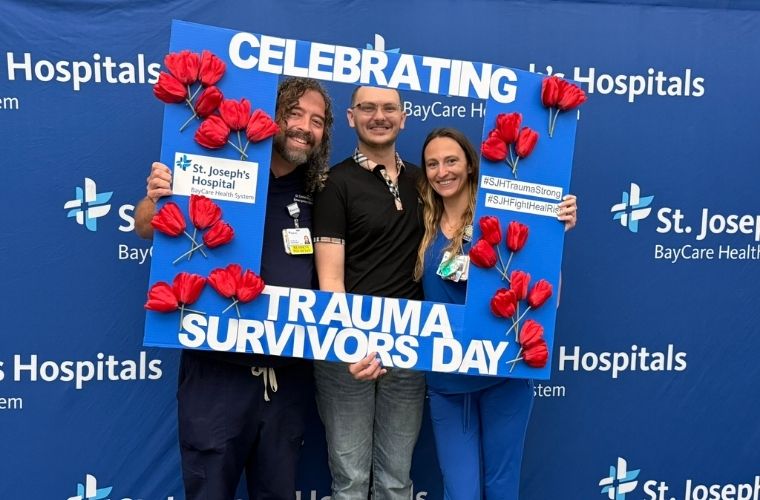 Three individuals smiling and holding a "Celebrating Trauma Survivors Day" frame decorated with red flowers. They are standing in front of a backdrop featuring the St. Joseph's Hospitals and BayCare Health System logos.