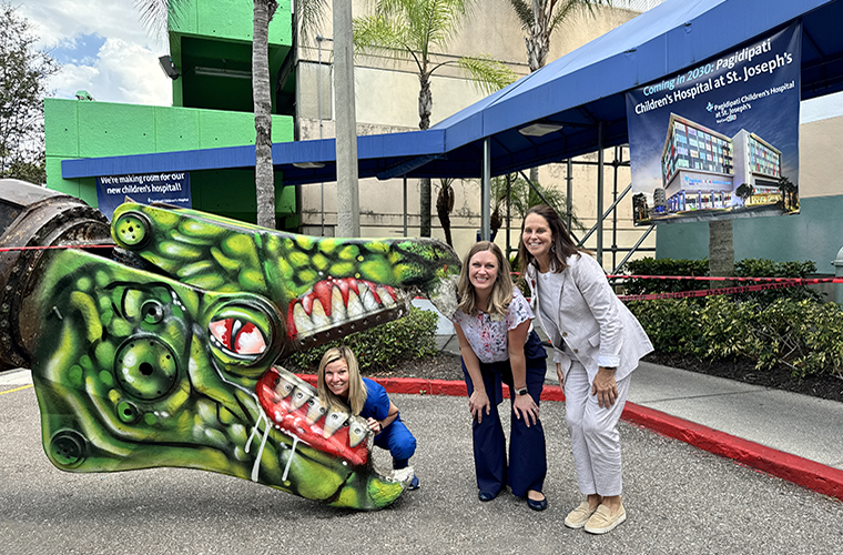 A woman wearing blue scrubs playfully puts her head in the jaws of a dinosaur painted excavator as two women, both wearing professional attire, crouch next to her. 