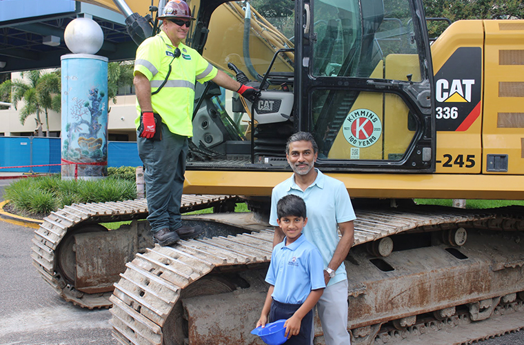 A man in neon construction vest stands on an excavator as another man and boy, both wearing blue shirts, stand in front of it.
