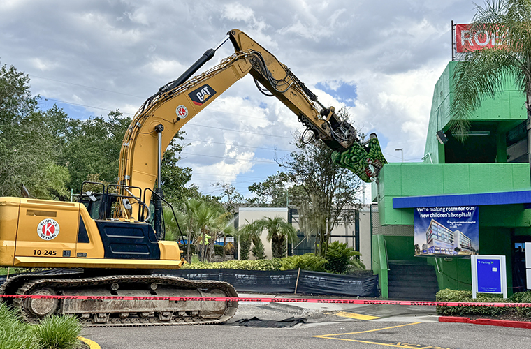 A dinosaur-painted excavator takes a bite out of a green hospital parking garage. The body of the excavator is yellow. There are trees in the background.