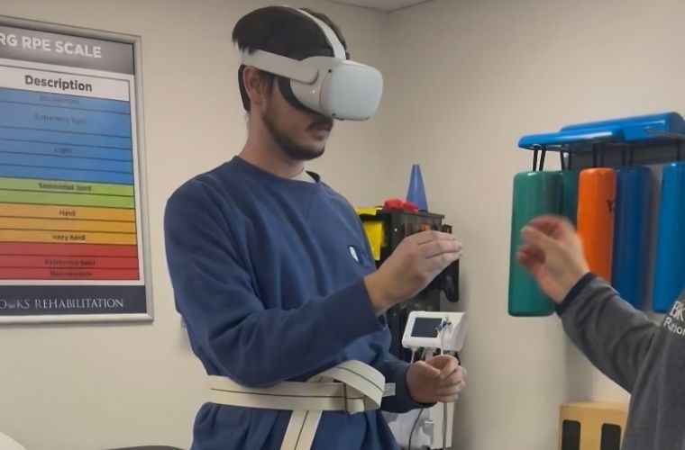 A young man stands with a virtual reality headset on moving his hands with the help of a rehab specialist.