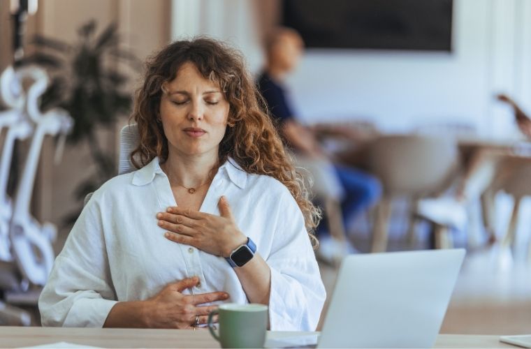 A young woman sits with a laptop and mug. She has one hand over her heart and one hand on her abdomen. Her eyes are closed as she focuses on mindful breathing techniques for relaxation and stress management.