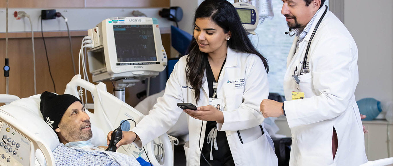 A faculty member assists a resident as she performs a medical test on an adult patient.