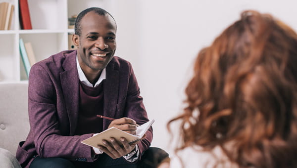 an African American therapist talking to a girl with reddish brown hair while holding a notepad and pencil