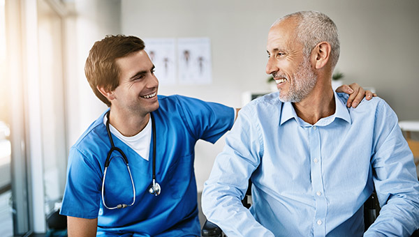 A young male nurse in blue scrubs putting his arm around an older male patient's shoulder, smiling and looking at each other
