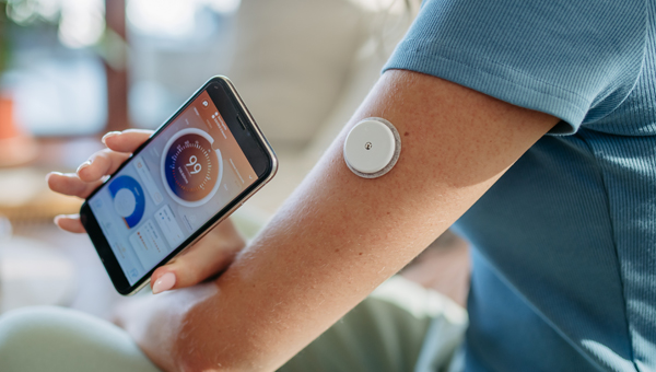 A patient holding a phone showing a connected medical device monitoring her blood sugar on the other arm. 