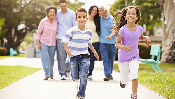 A young girl and boy running on a sidewalk while their parents and grandparents watch them.