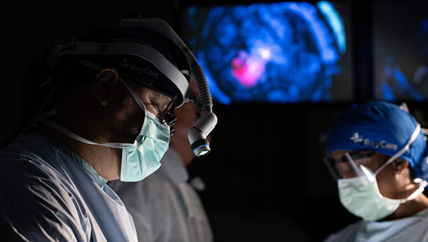 close up of a BayCare neurosurgeon wearing a mask surrounded by 2 other surgeons in dark lighting