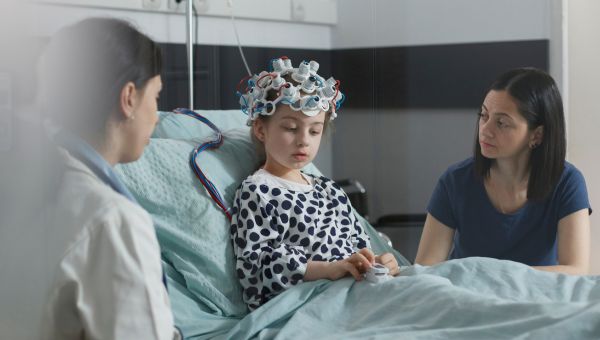 A mother and her daughter, who is in a hospital bed and wearing a device on her head, are meeting with their female neurologist. 