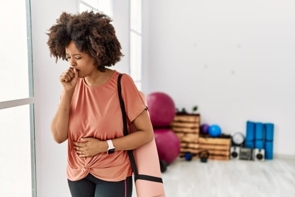 African american woman with afro hair holding yoga mat at pilates room feeling unwell and coughing.