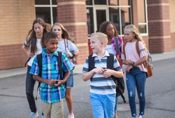 A diverse group of elementary aged kids walk away from a school and talk with each other.